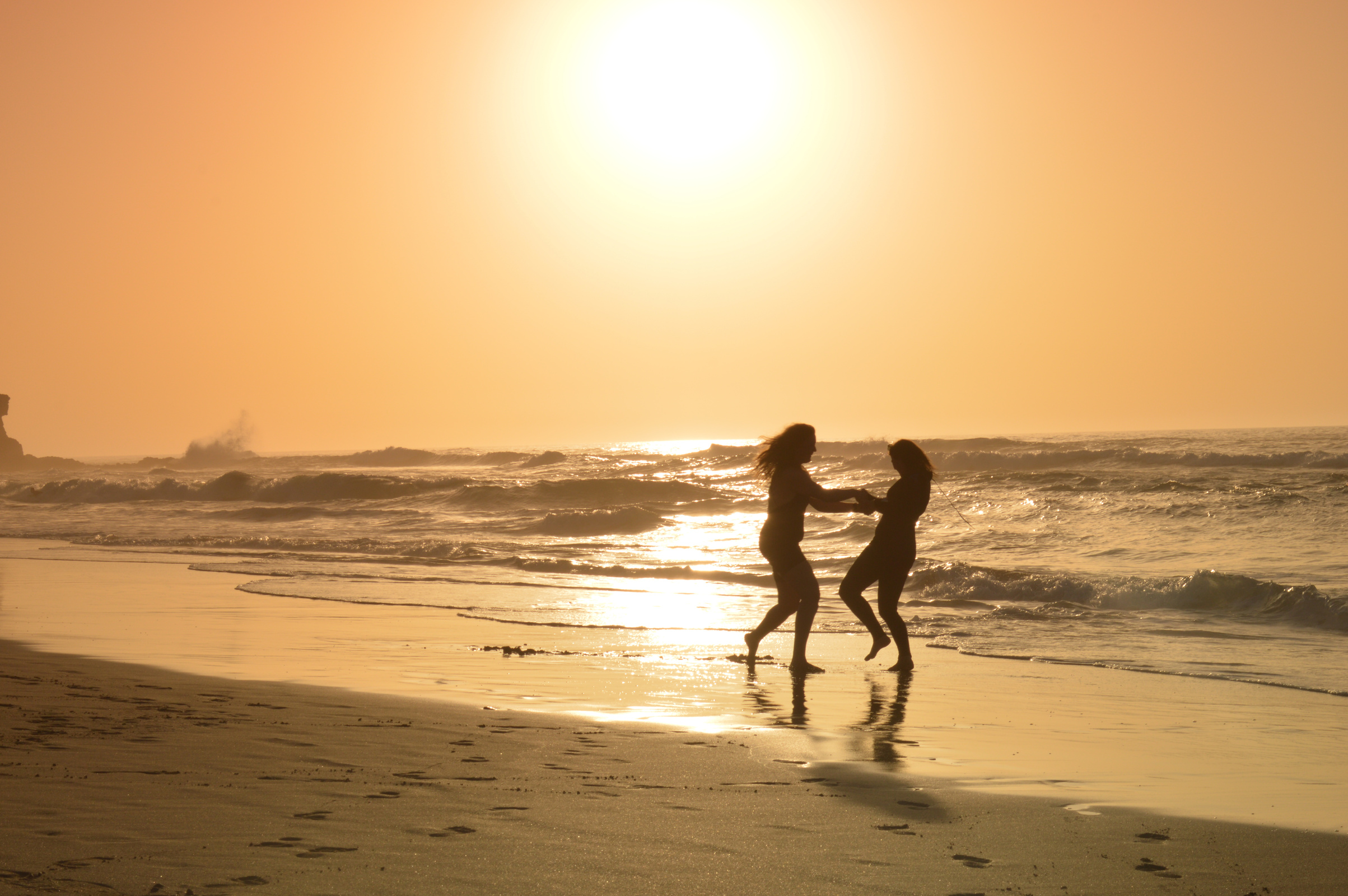 Background image: Two women playing holding hands at sunset on a beach.